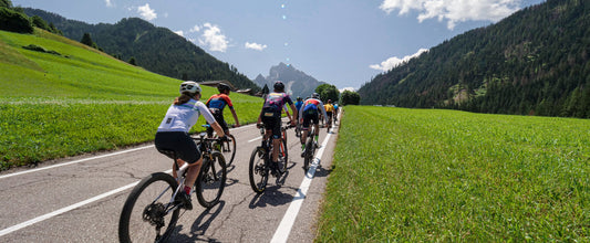 Participantes de Dolomiti SuperBike atravesando los verdes prados de Trentino. Al fondo de la imagen se ven las montañas de los Dolomitas.