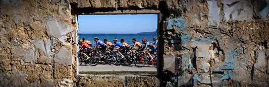 Pared de rocas con una abertura en forma de ventana tras la que se puede ver el pelotón ciclista y el mar.