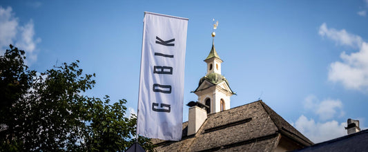 Bandera de color claro con el logo de la marca en tono oscuro ondeando. Al fondo, se ve el campanario de una iglesia y un árbol.